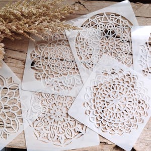 May include: Five white plastic stencils featuring intricate mandala designs. The stencils are arranged on a wooden surface.