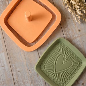 May include: A square orange silicone mold with a central cylindrical post and a square sage green silicone mold featuring an embossed heart design surrounded by radiating lines and a border of small heart shapes along the inner edge.