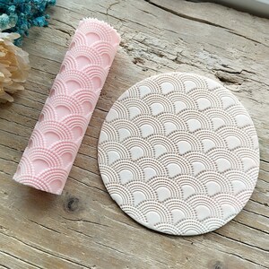 Könnte beinhalten: Ein rosa Nudelholz und eine weiße runde Silikonmatte mit einem sich wiederholenden Muster aus weißen Kreisen und Bögen. Beide Artikel sind zum Erstellen von dekorativen Mustern auf Fondant oder Teig gedacht.
