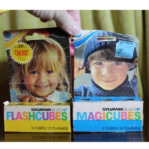 May include: Two Sylvania Blue Dot packaging boxes for photography. The left box is yellow, labeled Flashcubes, 3 cubes/12 flashes, featuring a child. The right box is blue, labeled Magicubes, 3 cubes/12 flashes, featuring a child in a blue hat.