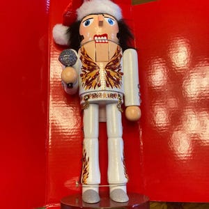 May include: A wooden Christmas nutcracker resembling Elvis Presley, dressed in a Santa hat and a white jumpsuit with gold and red detailing. The figure holds a silver microphone and stands on a round base with the name "ELVIS" inscribed.