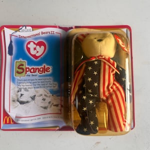May include: A Spangle the Bear Beanie Baby, part of the International Bears II collection, in its original packaging. The bear is dressed in a patriotic outfit with red and white stripes and a navy blue fabric with white stars. The packaging includes the Ty heart logo.