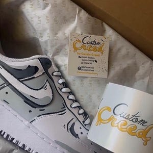 Op de afbeelding: Op maat gemaakte witte sneakers met een zwart en grijs camouflagepatroon. De sneakers zitten in een doos met een visitekaartje waarop "Custom Creed by Cassie Creed" staat.
