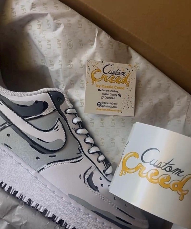 Op de afbeelding: Op maat gemaakte witte sneakers met een zwart en grijs camouflagepatroon. De sneakers zitten in een doos met een visitekaartje waarop "Custom Creed by Cassie Creed" staat.