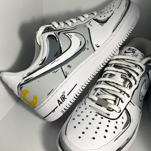 Op de afbeelding: Een paar witte sneakers met zwarte en grijze cartoon-achtige ontwerpen. De sneakers hebben gele accenten en de tekst "AIR" aan de zijkant.