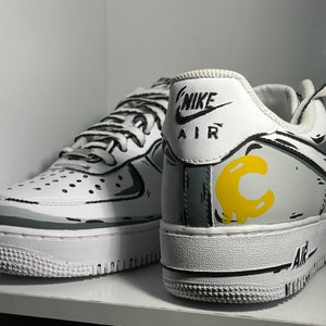 Op de afbeelding: Een paar witte Nike Air Force 1 sneakers met zwarte en grijze cartoon-achtige ontwerpen. De rechter schoen heeft een gele "C" aan de zijkant. De schoenen hebben witte veters en een zwarte Nike swoosh.