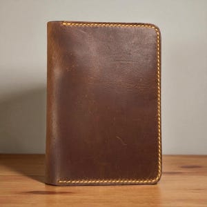 Puede incluir: Una cartera de cuero marrón con un borde cosido. La cartera es rectangular y se apoya sobre una superficie de madera. La costura es de color amarillo contrastante. El cuero tiene una veta visible.