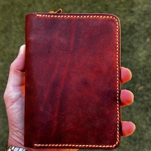 May include: A rectangular, deep burgundy leather wallet featuring a smooth, matte finish and visible grain texture. The edges are secured with thick, light-colored stitching that creates a distinct border around the entire perimeter of the wallet.