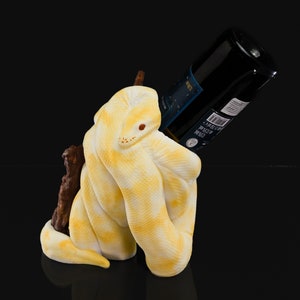 May include: A yellow and white ceramic wine bottle holder shaped like a snake. The bottle holder is holding a dark glass bottle of wine.
