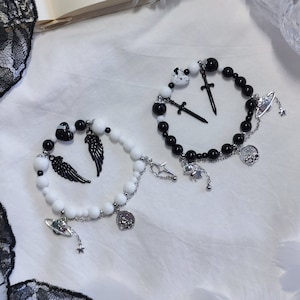 Puede incluir: Dos pulseras con cuentas blancas y negras y dijes plateados. Una pulsera tiene un dije plateado de un par de alas, un planeta y una estrella. La otra pulsera tiene un dije plateado de una espada, una cruz y una luna creciente.