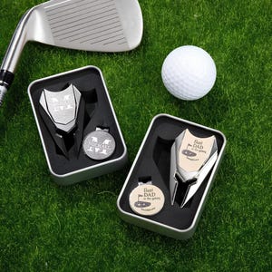 Könnte beinhalten: Zwei silberne Golf-Divot-Werkzeuge in Geschenkboxen. Eine Box enthält einen silbernen Golfball mit dem Text "Best Dad in the Galaxy" und die andere einen silbernen Golfball mit dem Text "Michael".
