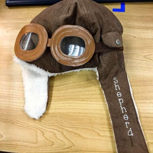 以下が含まれることがあります： 白いフェイクファーのトリムとゴーグルが付いた、茶色のフェイクレザーのパイロットハット。帽子には「SHEPHERD」という名前が側面に刺繍されています。