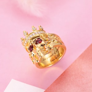 May include: A gold ring shaped like a crown with a skull motif, featuring small clear stones along the top edge and two deep red round stones set as eyes within the skull. The interior band is engraved with cursive text.