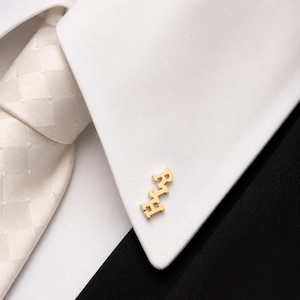 Pode incluir: Abotoaduras douradas com as iniciais "RJH" na manga de uma camisa branca. Uma gravata creme com um padrão subtil é visível no fundo, juntamente com um casaco preto.