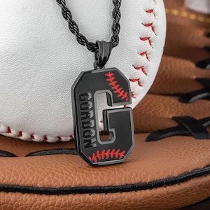 Op de afbeelding: Zwarte ketting met een hanger in de vorm van de letter "G" en de naam "GORDON" in het wit. De hanger heeft rode stikseldetails, die lijken op een honkbal. De ketting wordt getoond op een honkbalhandschoen en een honkbal.