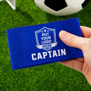 Może przedstawiać: Niebieska opaska kapitańska z białym tekstem i obszarem logo. Opaska zawiera słowo "CAPTAIN" dużymi białymi literami. Obszar logo zawiera tekst "PUT YOUR LOGO HERE". Piłka nożna i bramka są w tle.