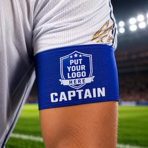 Könnte beinhalten: Eine blaue Kapitänsbinde mit dem Text "PUT YOUR LOGO HERE" und "CAPTAIN" in Weiß. Die Armbinde wird auf einem weißen und goldenen Sporttrikot getragen. Der Hintergrund zeigt ein Sportfeld.