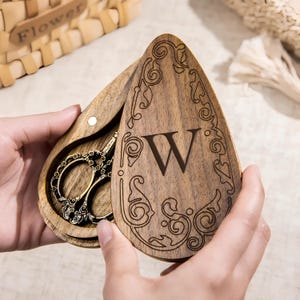 Peut inclure: Une boîte en bois avec un motif floral gravé et la lettre "W" sur le couvercle. La boîte est ouverte, révélant une paire de ciseaux de style antique à l'intérieur.