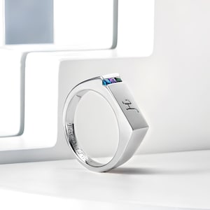 Op de afbeelding: Een zilveren ring met een rechthoekige band gegraveerd met de tekst "Best Dad Ever". De band is bezet met vier kleine edelstenen in een rij: blauw, groen, paars en blauw.
