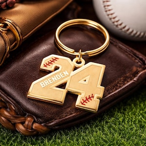 Könnte beinhalten: Goldfarbener Schlüsselanhänger mit der Zahl 24 und dem eingravierten Namen "Brenden". Der Schlüsselanhänger im Baseball-Design hat rote Nahtdetails. Er ist an einem goldenen Ring befestigt. Der Hintergrund umfasst einen Baseballhandschuh und einen Baseball.