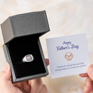 Può includere: Un anello d'argento con una pietra blu e la lettera "M" incisa sulla parte superiore. L'anello è in una scatola grigia con un biglietto che dice "Buon giorno del padre" e "Grazie per essere il miglior papà e nonno che potremmo chiedere".