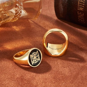 May include: Two gold rings. One ring has a black circular face with a geometric design. The other ring is a simple band with the inscription "Always and Forever" inside. The rings are on a brown surface.