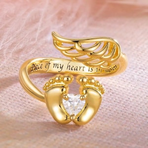 Könnte beinhalten: Goldfarbener Ring mit einem Paar Babyfüßen, die einen herzförmigen Edelstein umarmen. Der Ring hat ein Flügeldesign und die Inschrift "Piece of my heart is in heaven."