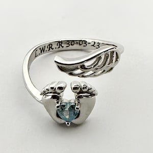 May include: A silver-colored ring with a heart-shaped blue gemstone held by two hands. The ring has a wing-like design and engraved text: L.W.R.R 30-03-23. The ring is set against a white background.