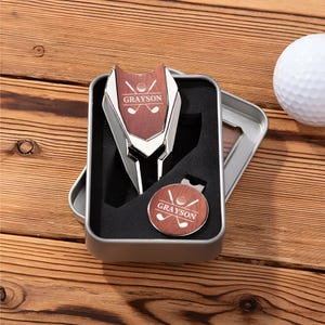 May include: A silver golf ball marker and a silver golf divot tool with a woodgrain design. The tools are in a silver box with a black interior. The tools are personalized with the name "Grayson" and a golf ball and clubs graphic.