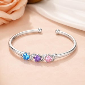 Pode incluir: Uma pulseira de punho aberta prateada com quatro pedras preciosas coloridas. As pedras são azuis, roxas e rosas, com os nomes "Lucas", "Emma" e "Olivia" gravados. A pulseira tem um design de coração.