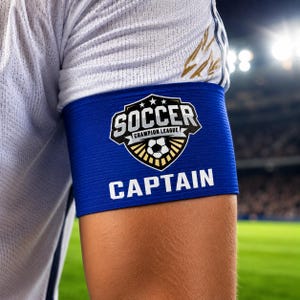 May include: A blue soccer captain's armband with the words "SOCCER CHAMPION LEAGUE" and "CAPTAIN" in white. The armband is worn on the upper arm of a person wearing a white soccer jersey.