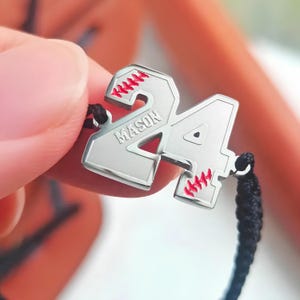 May include: A silver baseball charm with the number 24 and the name "MASON" engraved on it. The charm is attached to a black cord bracelet.