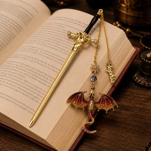 K&ouml;nnte beinhalten: Ein goldfarbener, schwertf&ouml;rmiger Lesezeichen mit schwarzem Griff liegt auf einem offenen Buch. Ein Drachenanh&auml;nger mit roten und goldenen Fl&uuml;geln und ein Sternenanh&auml;nger h&auml;ngen an einer Kette. Der Name "Sophia" steht auf einem goldenen Anh&auml;nger.