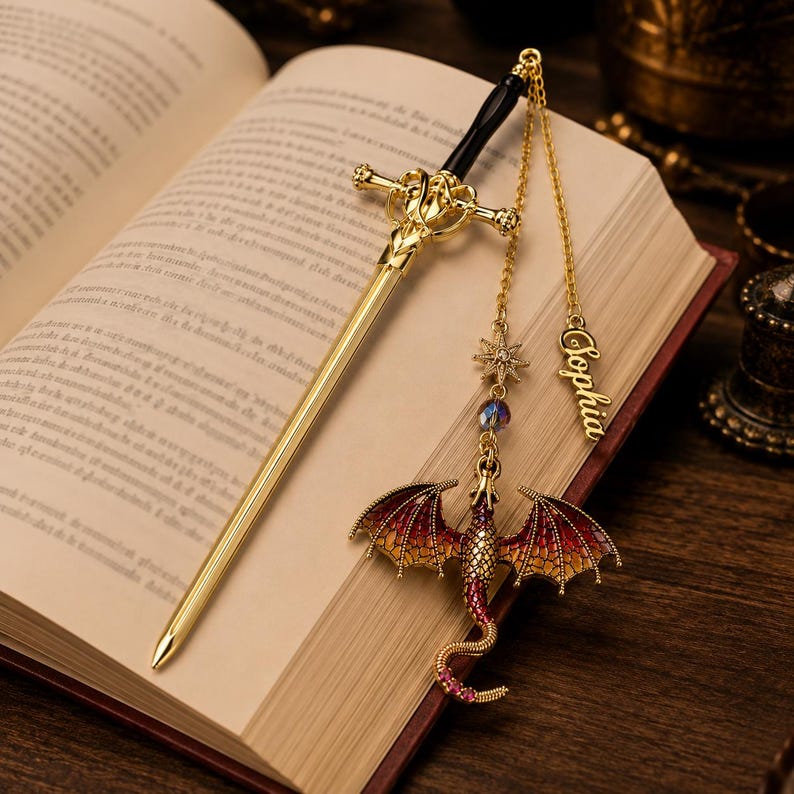 K&ouml;nnte beinhalten: Ein goldfarbener, schwertf&ouml;rmiger Lesezeichen mit schwarzem Griff liegt auf einem offenen Buch. Ein Drachenanh&auml;nger mit roten und goldenen Fl&uuml;geln und ein Sternenanh&auml;nger h&auml;ngen an einer Kette. Der Name "Sophia" steht auf einem goldenen Anh&auml;nger.