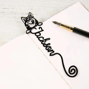 May include: Black metal bookmark featuring a husky dog design with the name "Jackson" in a cursive font. The bookmark has a decorative swirl at the bottom and is placed on a white page next to a pen.