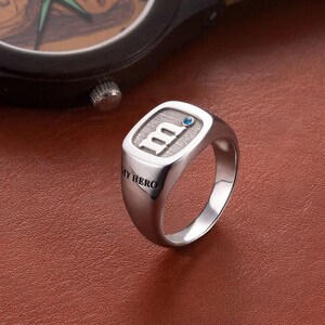 Può includere: Un anello d'argento con una faccia rettangolare con la lettera "m" e una piccola gemma blu. Le parole "MY HERO" sono incise sulla fascia dell'anello. L'anello è esposto su una superficie marrone.