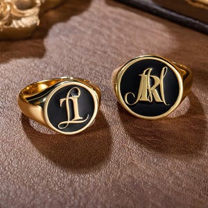 May include: Two gold signet rings with black enamel. One ring features the initials "LI" and the other ring features the initials "MR".