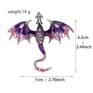 K&ouml;nnte beinhalten: Eine lila-silberne Drachenbrosche mit rosa und lila Akzenten. Der Drache hat ausgebreitete Fl&uuml;gel und ist mit Strasssteinen verziert. Die Brosche ist 7 cm lang und 6,2 cm breit.