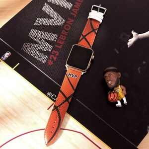 Puede incluir: Un Apple Watch con caja dorada y correa con temática de baloncesto. La correa naranja presenta líneas negras y la palabra "MVP". También hay una pequeña figura de un jugador de baloncesto.