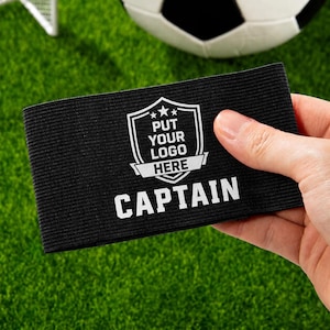 Könnte beinhalten: Schwarze elastische Armbinde mit dem Wort "CAPTAIN" in weißen Blockbuchstaben. Die Armbinde hat ein weißes Schilddesign mit dem Text "PUT YOUR LOGO HERE". Ein Fußball und grüner Rasen im Hintergrund.
