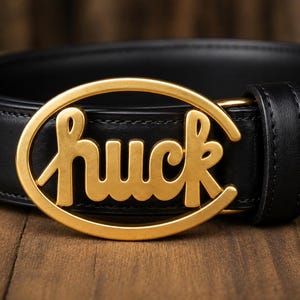 Peut inclure: Une ceinture en cuir noir dotée d'une boucle ovale dorée avec le mot huck écrit en écriture cursive stylisée. La ceinture comprend une attache à bouton-pression en métal doré assortie sur la sangle près de la boucle.