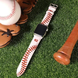 Könnte beinhalten: Weißes Lederarmband für Apple Watch mit einem Baseball-Steppdesign und der Nummer "42" darauf gedruckt. Das Armband liegt auf grünem Gras mit einem Baseballschläger und einem Handschuh im Hintergrund.