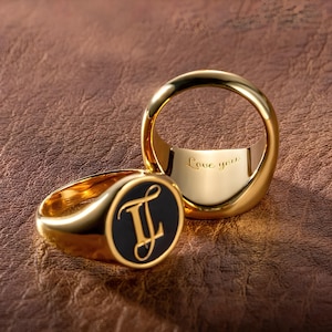 Può includere: Due anelli a sigillo in oro. Un anello ha un ovale nero con le iniziali "JL" in scrittura dorata. L'altro anello ha l'iscrizione "Love you" incisa all'interno.