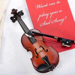 May include: A miniature wooden violin with a black bow and strings. The violin is resting on a white surface next to a red card with the text "Want me to play you a sad song?"