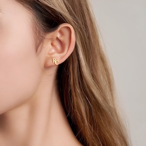 Peut inclure: Gros plan sur l'oreille d'une personne avec une boucle d'oreille initiale en or. La boucle d'oreille est un modèle simple avec les lettres "RC" dans une police classique.