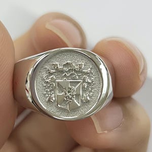 May include: A silver signet ring with a detailed crest design. The ring has a polished, reflective surface and a flat, circular face with an embossed heraldic emblem. The ring is held by a person.