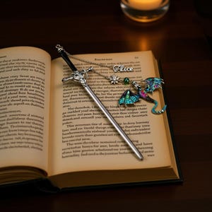 K&ouml;nnte beinhalten: Ein silberner, schwertf&ouml;rmiger Lesezeichen und eine Halskette mit einem Drachenanh&auml;nger und dem Namen "Alice" liegen auf einem offenen Buch. Der Drachenanh&auml;nger hat blaue, gr&uuml;ne und rosa Akzente. Im Hintergrund brennt eine Kerze.