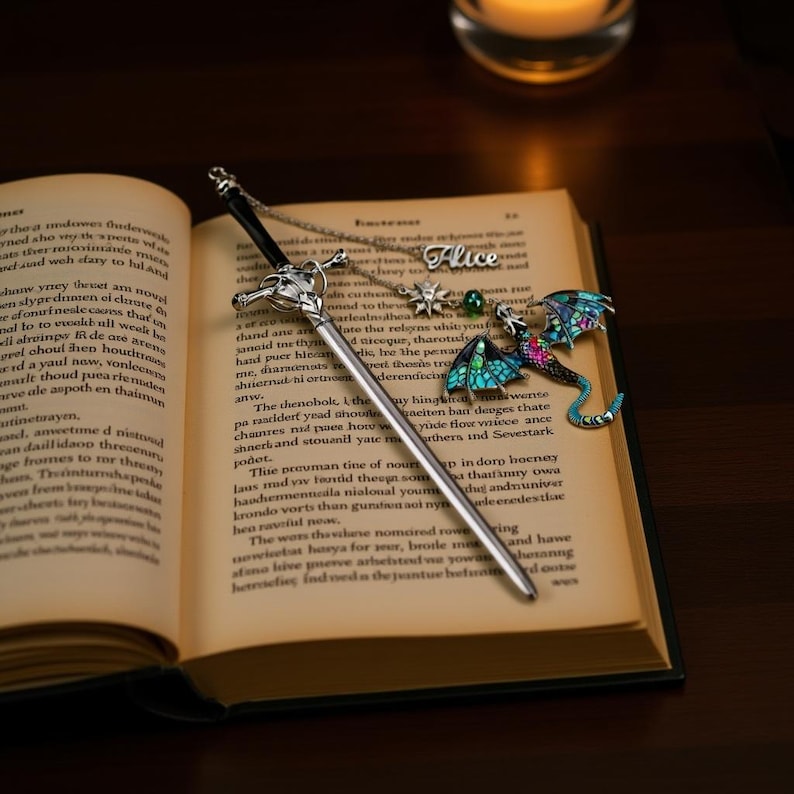 K&ouml;nnte beinhalten: Ein silberner, schwertf&ouml;rmiger Lesezeichen und eine Halskette mit einem Drachenanh&auml;nger und dem Namen "Alice" liegen auf einem offenen Buch. Der Drachenanh&auml;nger hat blaue, gr&uuml;ne und rosa Akzente. Im Hintergrund brennt eine Kerze.