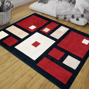 May include: A rectangular rug with a geometric pattern. It has a dark blue border and features red, cream, and white rectangular and square shapes. The rug is placed on a wooden floor.