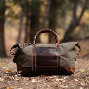 May include: A green canvas and brown leather duffel bag with a top handle and shoulder strap. The bag has a large brown leather panel on the front and bottom, with brass-colored hardware. The bag is sitting on a bed of leaves.